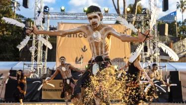A dancer with feathers in both handskicks up sand during a performance for DanceRites.